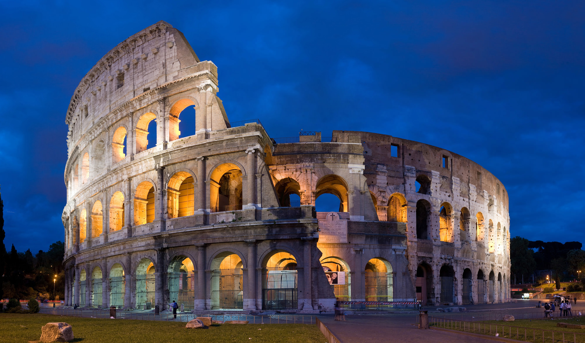 The Colosseum in Rome lit at dusk, with arches and exterior walls visible.